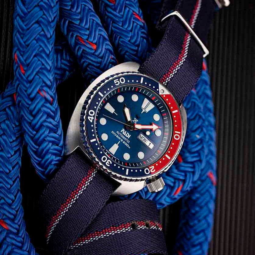 The Engraved Stripe in Red, White, and Blue watch by RSM features a blue dial with white indices, a blue and red bezel, and navy strap on a textured background.