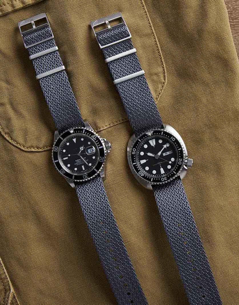 Two watches with black dials and round bezels on khaki fabric, each featuring RSM Herringbone Twill (Field Gray) NATO straps with silver detailing.