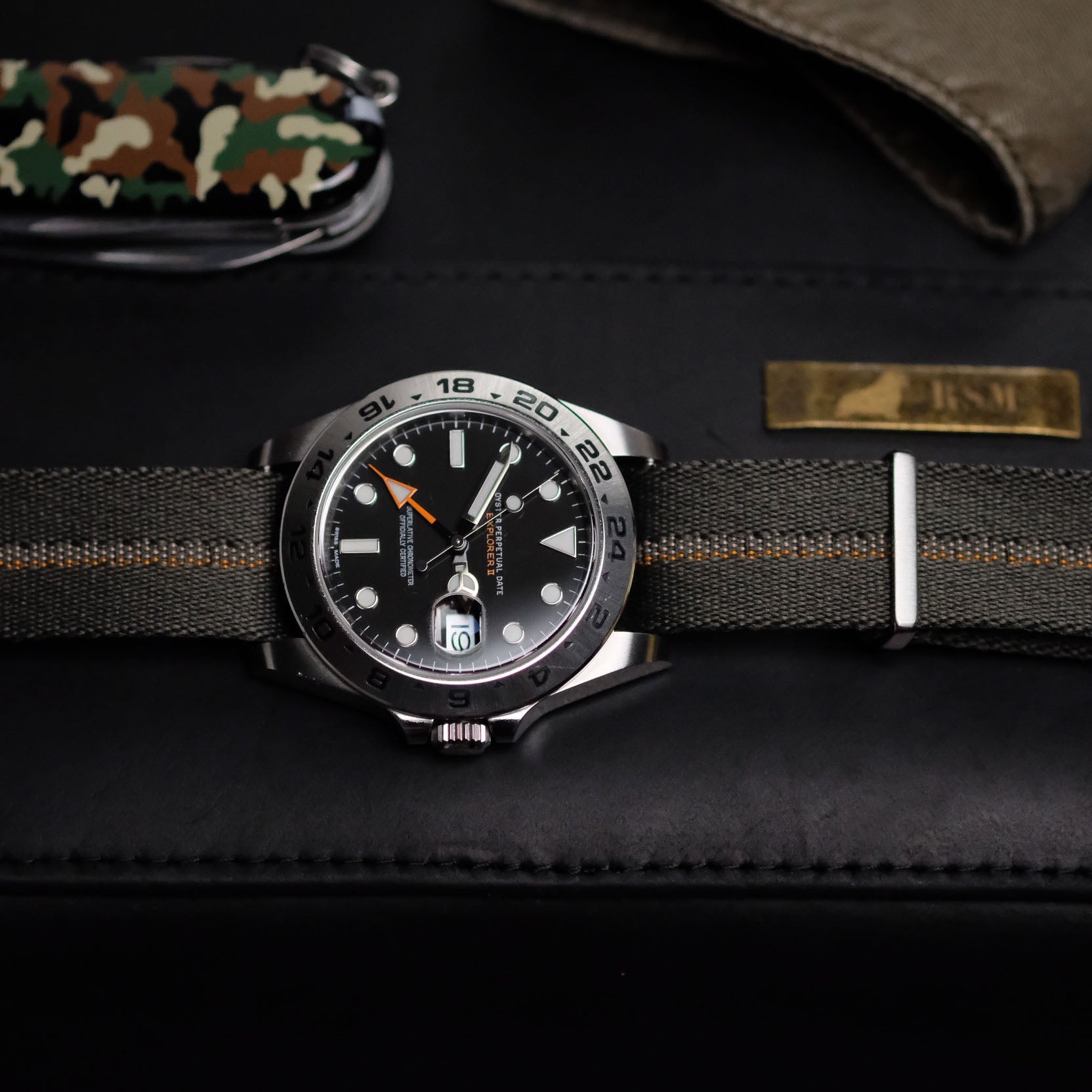 RSM Stripe Green, Beige, and Orange strap on a Rolex black-dial watch with orange details, placed on black with a Swiss army tool.